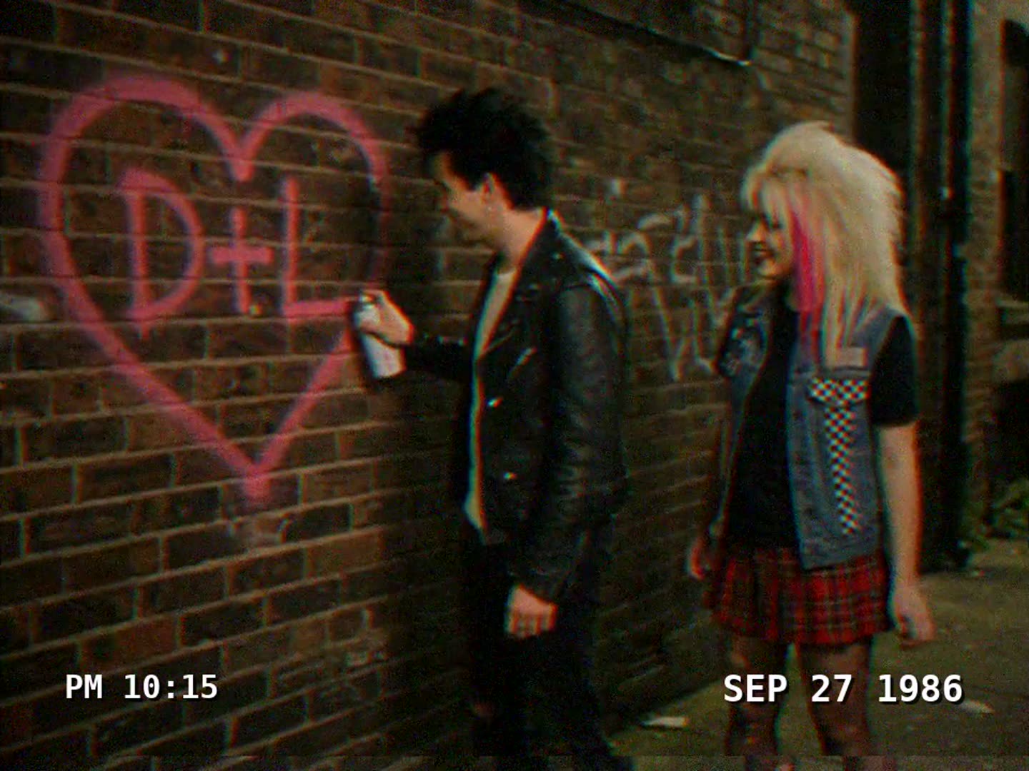 VHS home-video frame from 1986: a punk couple painting a heart on a brick wall
